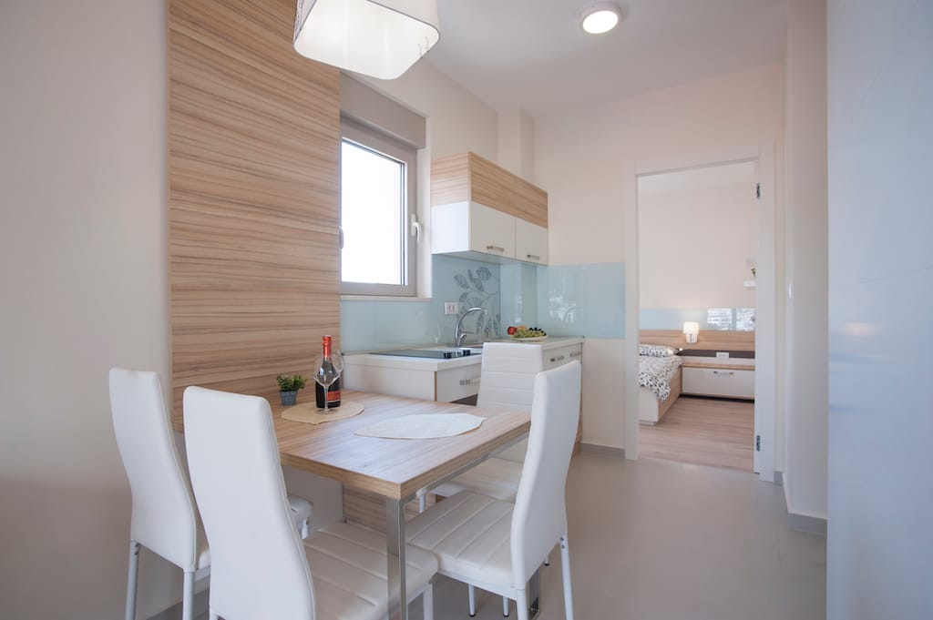 Savremena kuhinja-trpezarija u apartmanima Budve, Crna Gora: svetao nameštaj, trpezarijski deo i pogled na spavaću sobu.