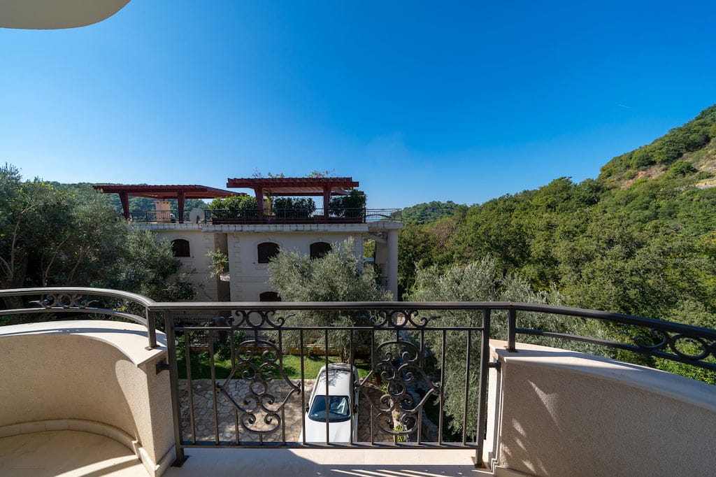 View from the apartment balcony in Petrovac, Montenegro: wrought-iron railings, a green garden, mountains, and a neighboring villa with a terrace.