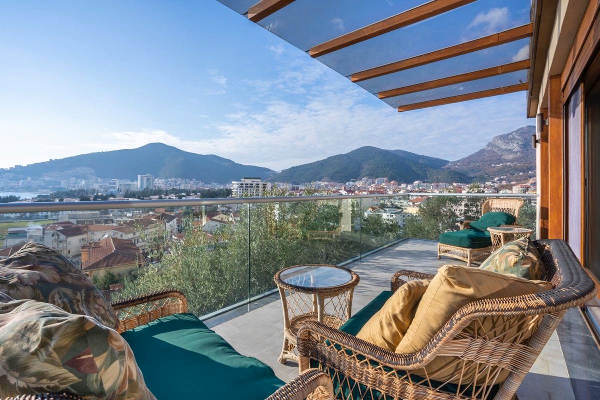 Terasa vile u Budvi, Crna Gora: pleteni nameštaj, staklena ograda i panoramski pogled na grad i planine.