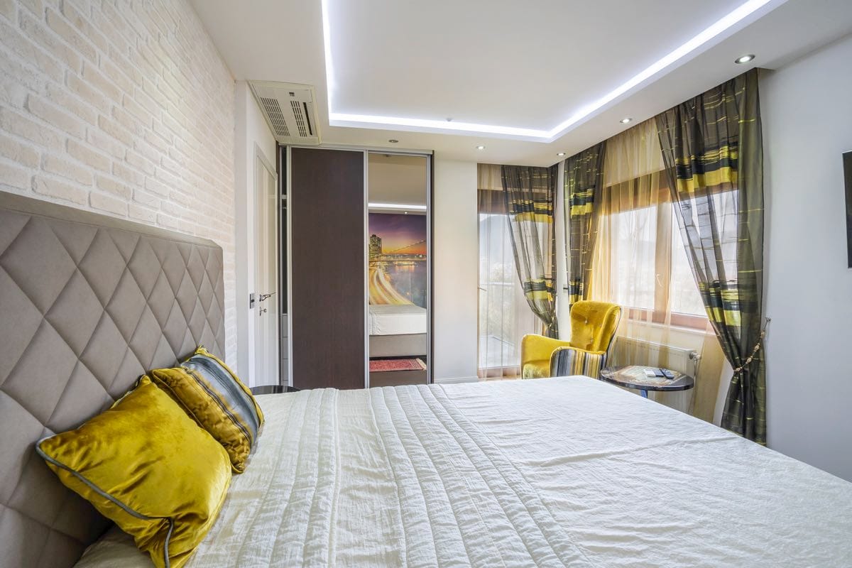 Savremena spavaća soba u vili u Budvi, Crna Gora: bračni krevet, zid od cigle, garderober i žuta fotelja.