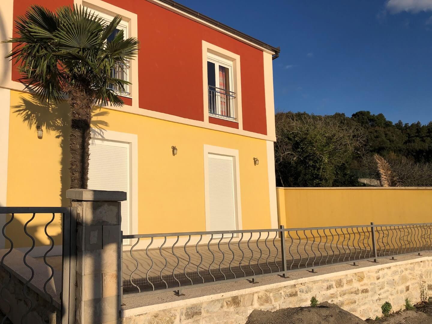 Fassade eines zweistöckigen Hauses in Herceg Novi, Montenegro: gelb-rote Wände, eine Palme und ein Metallzaun des Grundstücks.