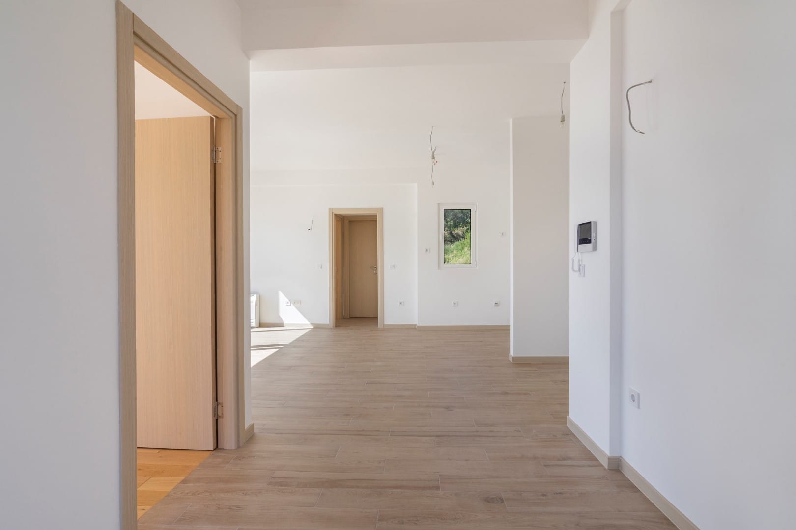 Svetao dnevni boravak u novom stanu u Tivtu, Crna Gora: beli zidovi, drveni pod, unutrašnja vrata i pogled na baštu.