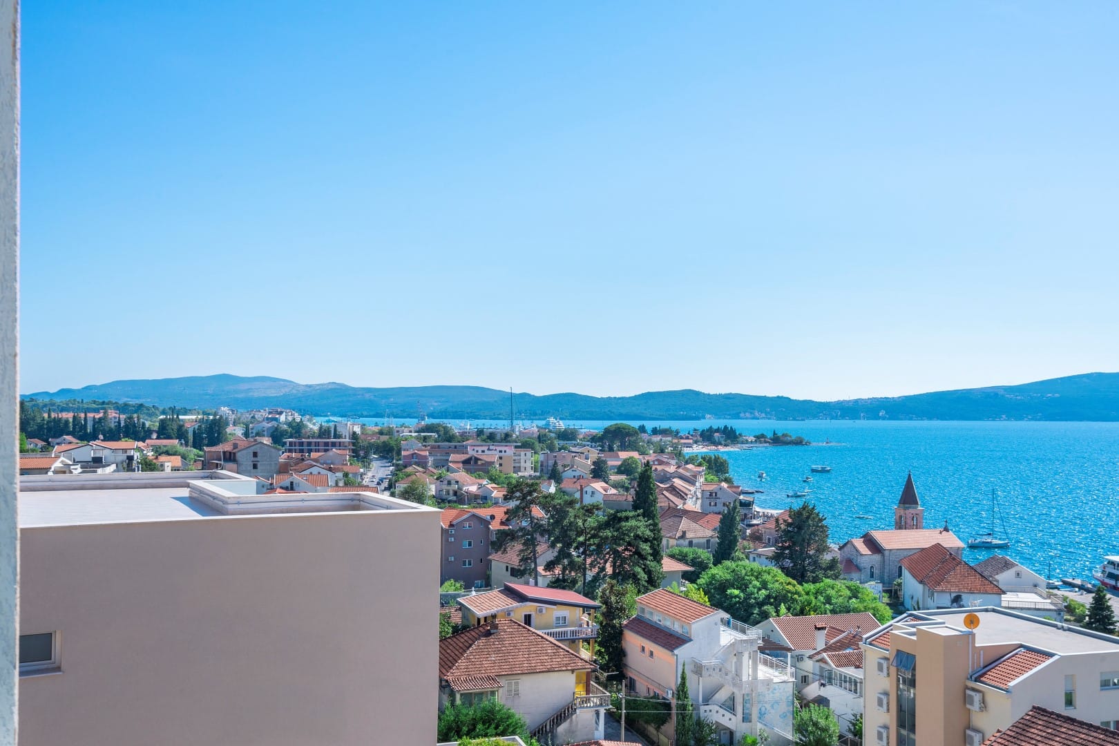 Panoramski pogled sa balkona stana u Tivtu, Crna Gora: morski zaliv, crveni krovovi kuća i planine na horizontu.