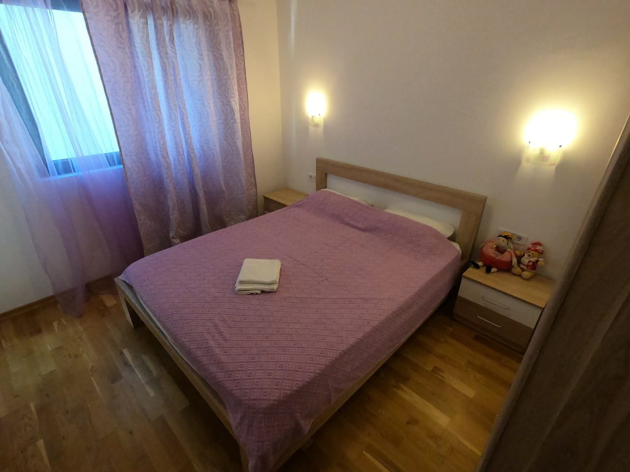 Udobna spavaća soba sa bračnim krevetom i parketom u stanu u Pržnu, Crna Gora. Svetao enterijer, zidne lampe.