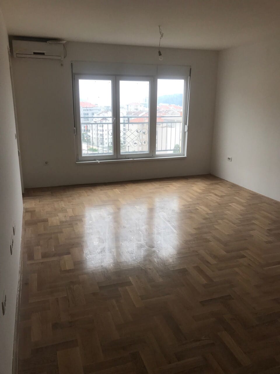 Svetla dnevna soba u stanu u Budvi, Crna Gora: parket, beli zidovi, klima-uređaj i pogled na grad kroz prozor.