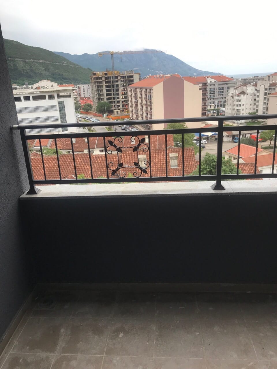 Pogled sa balkona stana u Budvi, Crna Gora: kovana ograda, gradska izgradnja i planinski pejzaž u pozadini.
