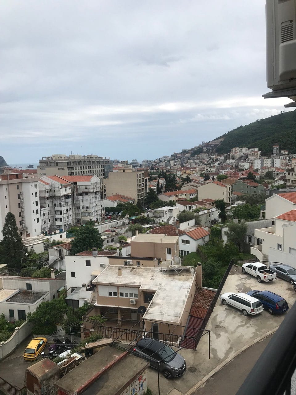 Pogled sa balkona stana u Budvi, Crna Gora: panorama grada, stambene zgrade, planine i parking pored zgrade.
