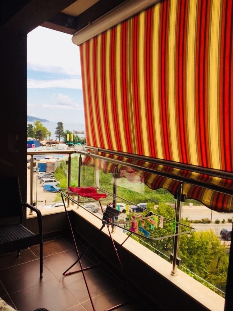 Balkon stana u Budvi, Crna Gora: prugasta tenda, staklena ograda i pogled na more i parking.