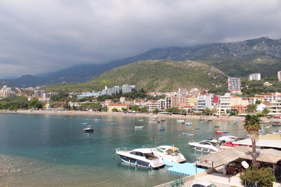 Pogled na more, plažu i jahte u Rafailovićima, Crna Gora: panorama letovališnog grada sa planinama i obalnom infrastrukturom.