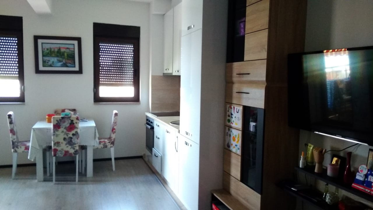 Dnevna soba sa kuhinjom u stanu u Budvi, Crna Gora: trpezarijski deo, svetao kuhinjski nameštaj i TV zona sa nameštajem.