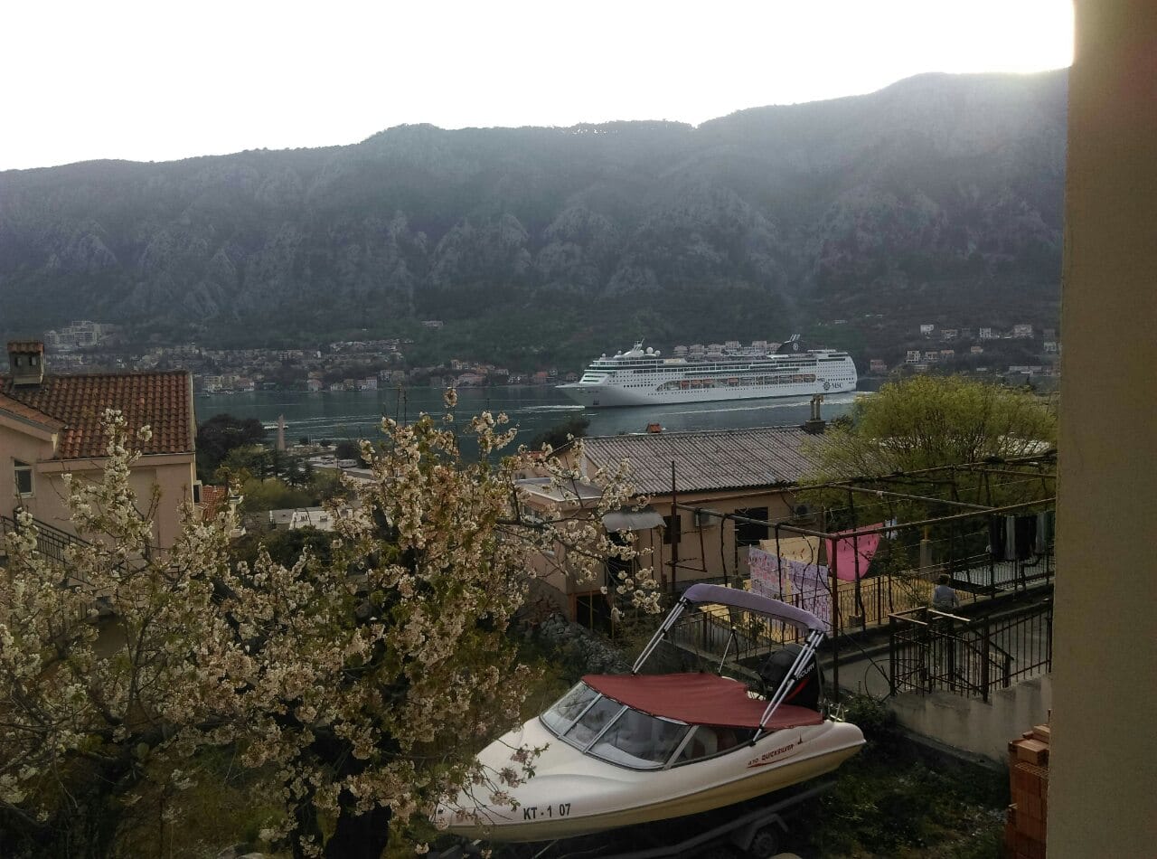 Pogled sa balkona stana u Kotoru, Crna Gora: panorama zaliva sa kruzerom, planinama i privatnim brodom na vezu.