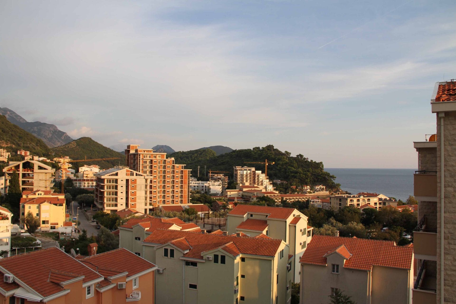 Panoramic view of Petrovac, Montenegro: residential buildings with tiled roofs, mountains, and the coastline.