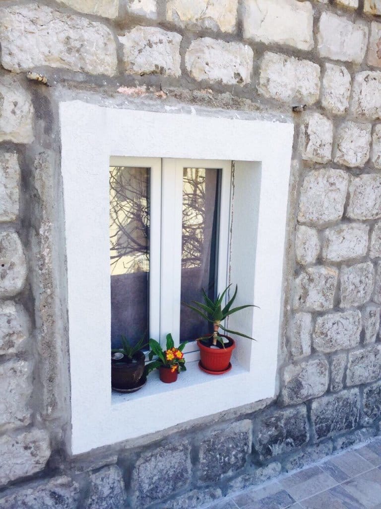 Beli plastični prozor u kamenoj zidu mini-hotela u Rafailovićima, Crna Gora, sa sobnim biljkama na prozorskoj dasci.