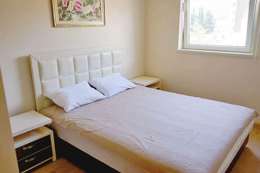 Helles Schlafzimmer mit Doppelbett in einer Wohnung in Budva, Montenegro: gepolstertes Kopfteil, Nachttische und Blick aus dem Fenster.