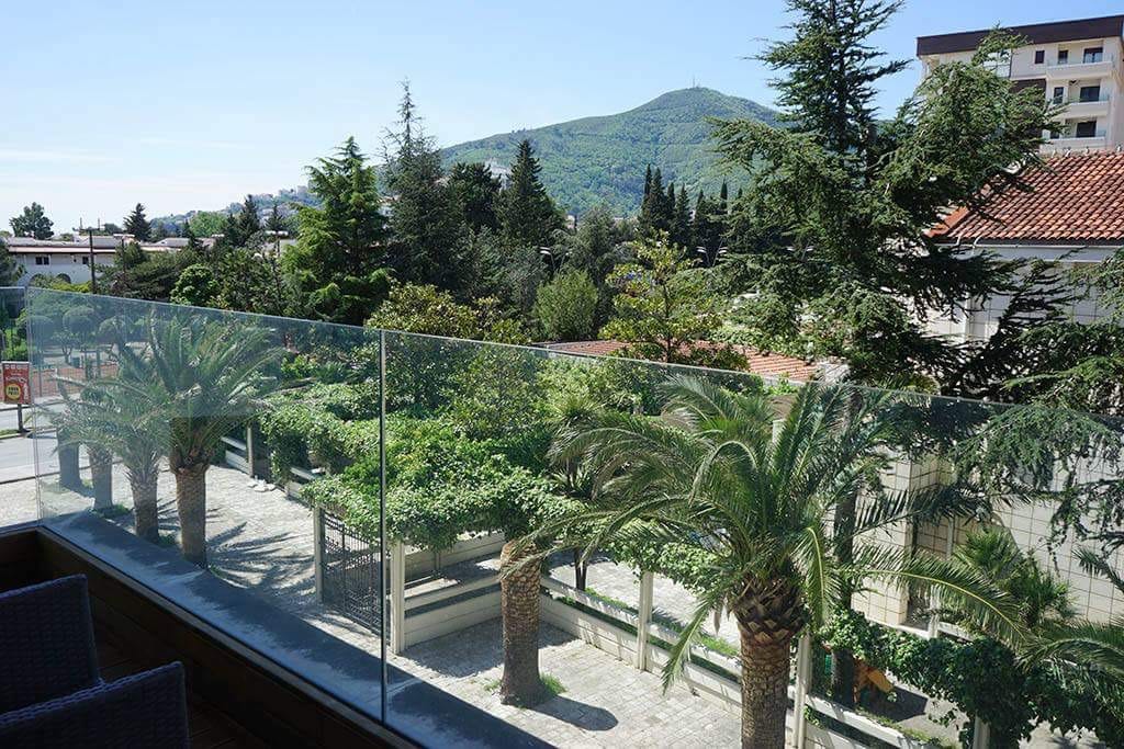 Blick vom Balkon der Wohnung in Budva, Montenegro: Glasgeländer, Palmen, grüner Garten und Berge im Hintergrund.