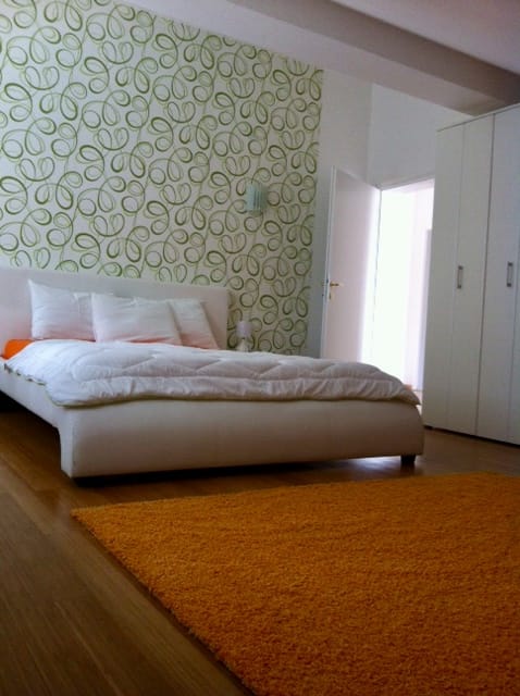 Schlafzimmer in einer Wohnung in Đenovići, Montenegro: weißes Bett, Akzentwand mit Muster, Parkett und ein leuchtend orangefarbener Teppich.