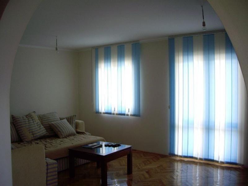 Svetla dnevna soba u kući u Sutomoru, Crna Gora: sofa, stočić za kafu, parket i vertikalne žaluzine.