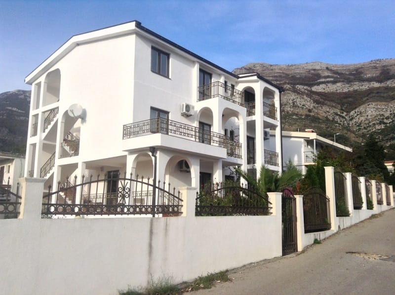 Trospratna bela kuća sa balkonima i kovanom ogradom u Sutomoru, Crna Gora, u pozadini planinski pejzaž.