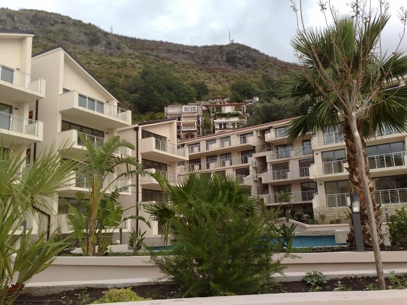 Moderner Wohnkomplex mit Pool und Palmen in Pržno, Montenegro: Blick auf die Fassaden der Wohnungen und die Berge.