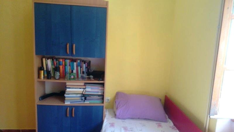 Chambre dans une maison à Dobrota, Monténégro : armoire bleue avec des livres, lit simple avec un coussin rose et murs jaunes.