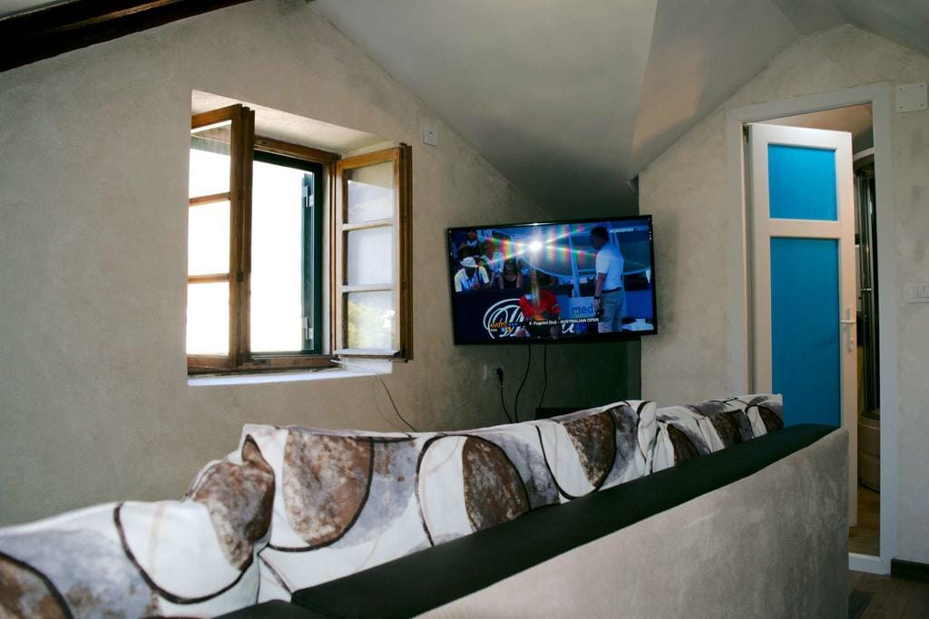 Salon dans une maison à Dobrota, Monténégro : canapé confortable, TV murale et fenêtre avec des murs aux finitions claires dans l’intérieur.