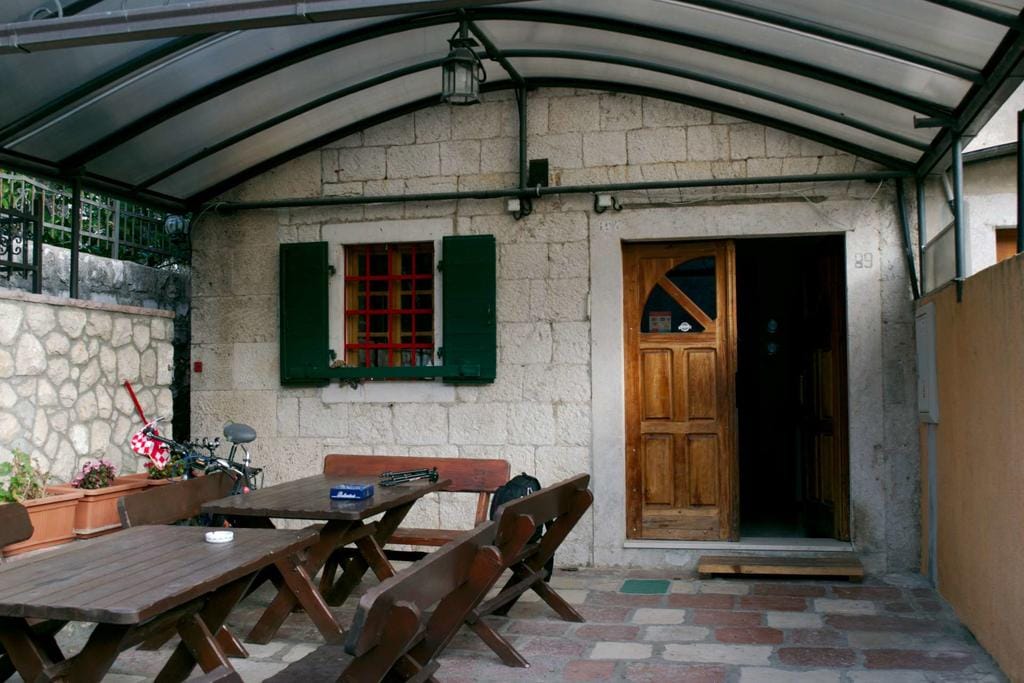 Maison en pierre à Dobrota, Monténégro : terrasse avec mobilier en bois, auvent et volets verts traditionnels.
