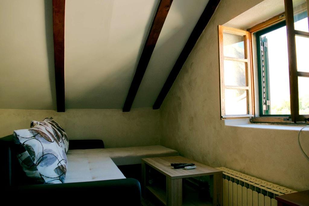 Salon chaleureux sous les combles d’une maison à Dobrota, Monténégro : canapé, poutres en bois au plafond et vue par la fenêtre.
