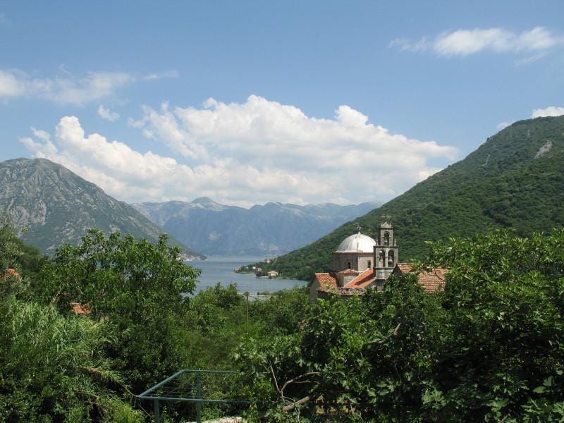 Vue panoramique sur la baie de Kotor et une ancienne église à Dobrota, Monténégro, entourée de montagnes et de verdure.