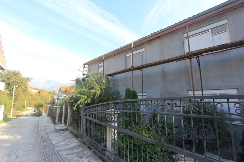 Maison à deux étages à Dobrota, Monténégro, avec façade en cours de rénovation et clôture en fer forgé sur fond de montagnes.