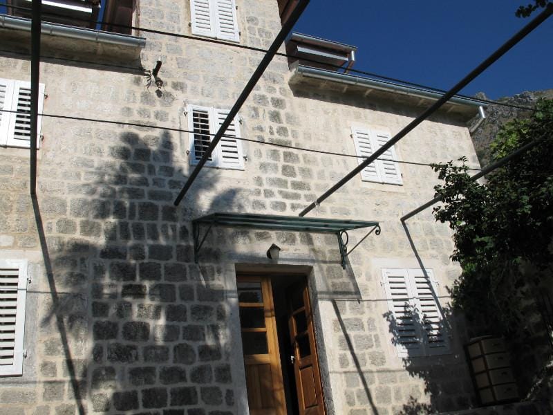 Façade d’une maison traditionnelle en pierre à Dobrota, Monténégro, avec volets blancs et terrasse sous auvent.