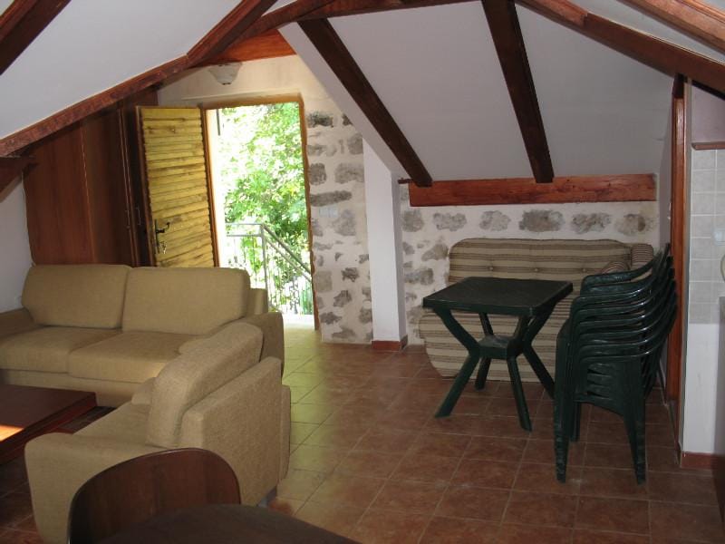Salon sous les combles d’une maison à Dobrota, Monténégro : murs en pierre, poutres en bois, mobilier moelleux et accès à la terrasse.