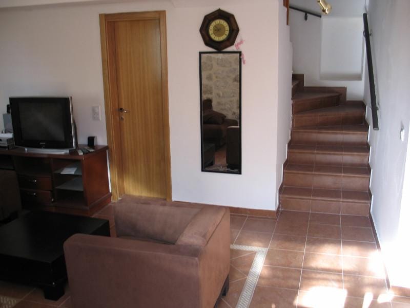 Salon dans une maison à Dobrota, Monténégro : fauteuil marron, télévision, porte en bois et escalier menant au deuxième étage.