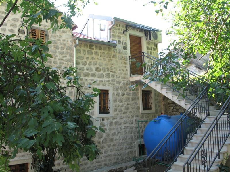 Façade d’une maison en pierre à Dobrota, Monténégro : escalier extérieur, balcon et réservoir d’eau dans un jardin verdoyant.