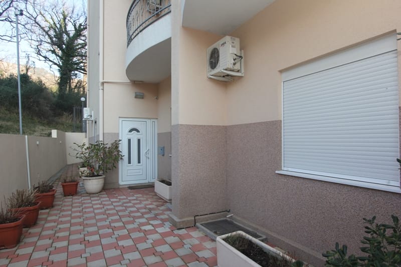 Ulaz u stan u Bečićima, Crna Gora: terasa sa pločicama, bela vrata, prozor sa roletnama i klima-uređaj na fasadi.