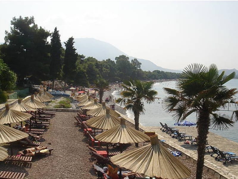 Beach with sun loungers and straw umbrellas by the sea in Shushan, Montenegro, with a view of the coastline and mountains.
