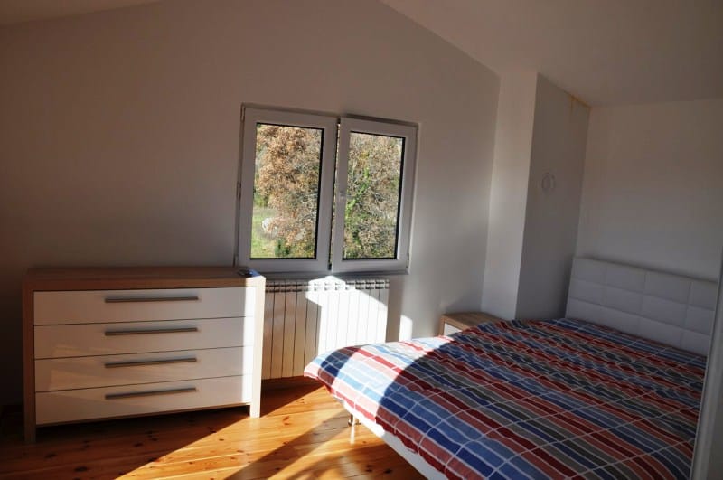 Svetla spavaća soba sa bračnim krevetom i komodom u kući na poluostrvu Luštica, Crna Gora, pogled na baštu iz prozora.