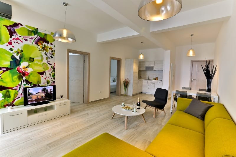 Moderna dnevna soba sa živopisnim dekorom i kuhinjom u stanu, Dobri Vodi, Crna Gora: žuti kauč, TV i svetao nameštaj.