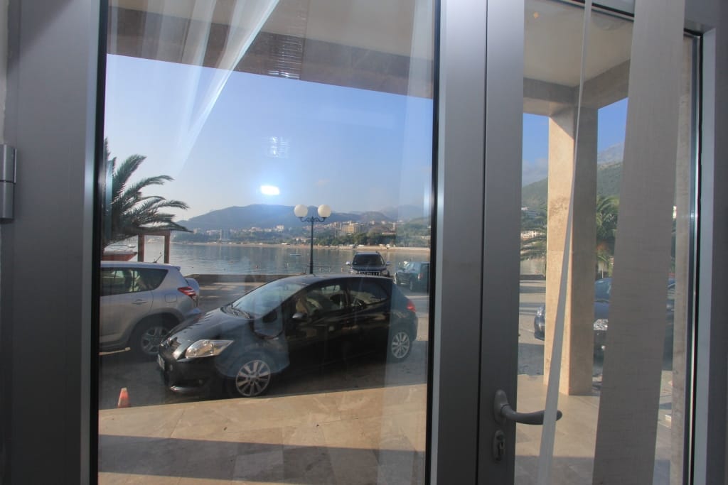 Pogled iz prozora poslovnog prostora na more, plažu i planine u Rafailovićima, Crna Gora, sa parkingom ispred ulaza.
