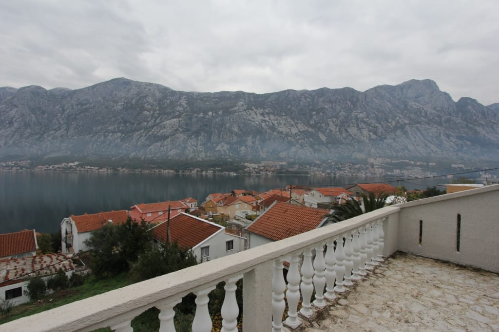 Widok z tarasu z balustradą na Zatokę Kotorską i góry w Prčanj, Czarnogóra: panorama morza i dachów domów z dachówki.