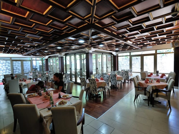 Wnętrze restauracji hotelowej w Petrovacu, Czarnogóra: drewniany sufit kasetonowy, panoramiczne okna i miękkie krzesła.