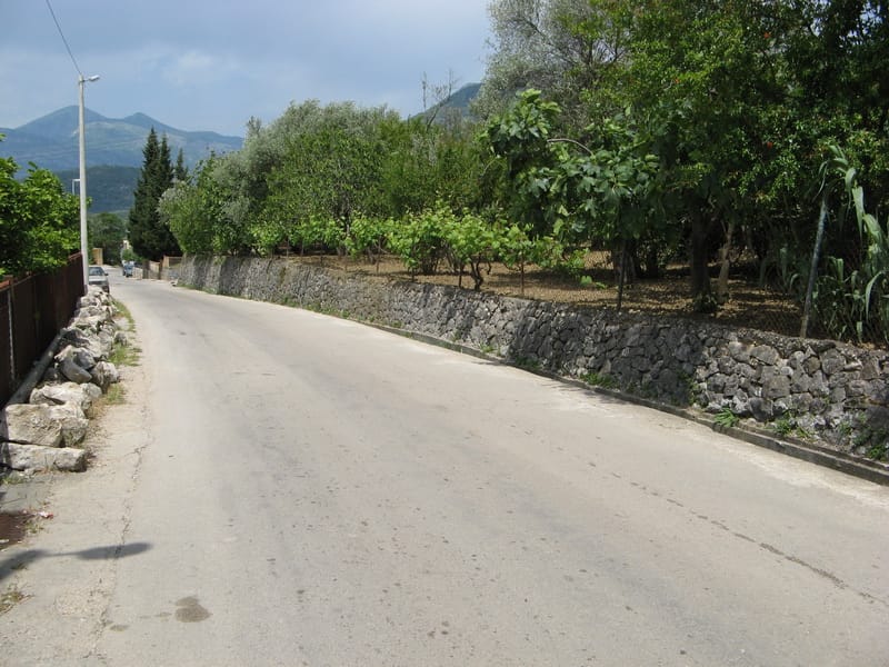 Asphaltierte Straße entlang des Grundstücks mit Steinmauer und Garten in Bar, Montenegro.