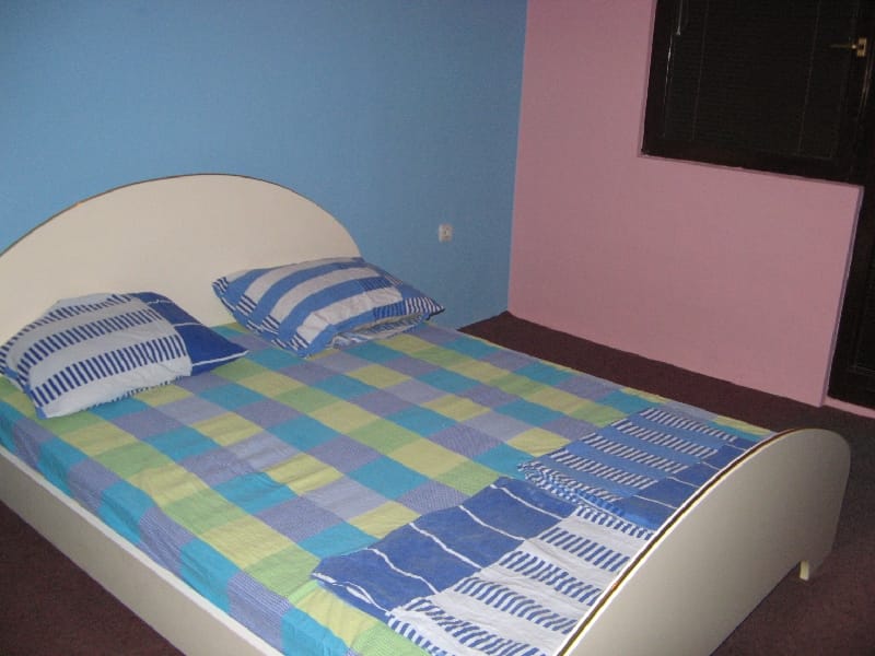 Spavaća soba sa bračnim krevetom u kući, Šušanj, Crna Gora: svetli zidovi, karirano posteljno rublje i tamni pod.
