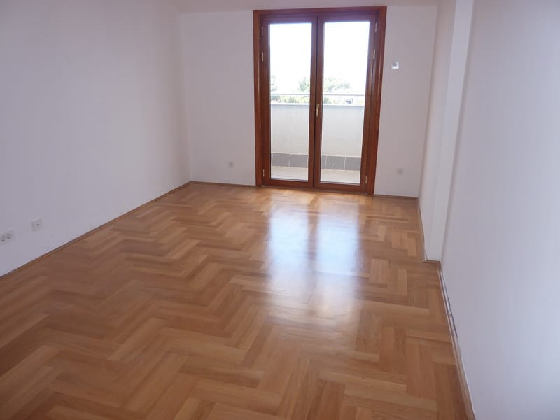 Svetao dnevni boravak sa parketom i izlazom na balkon u stanu u Tivtu, Crna Gora. Prazna soba sa belim zidovima.