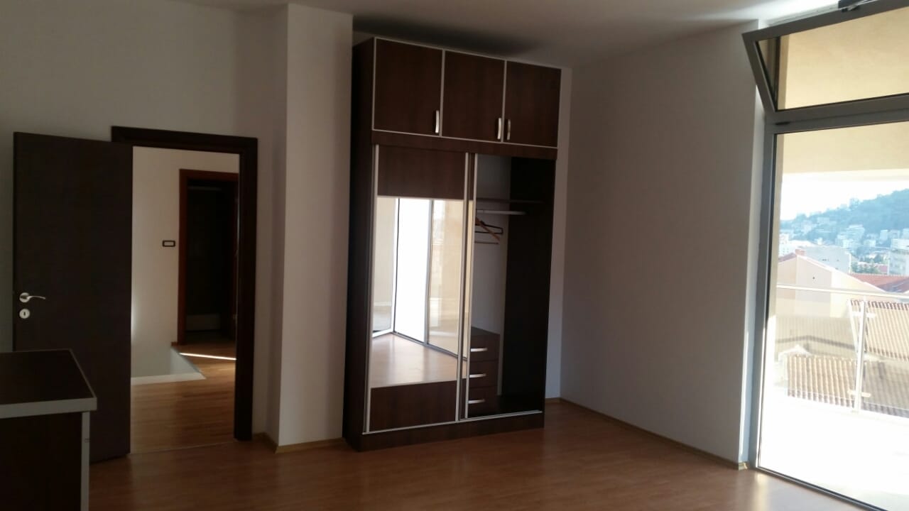 Spavaća soba u stanu u Budvi, Crna Gora: tamni ugradni ormar sa ogledalom, parketni pod i izlaz na balkon s pogledom na grad.