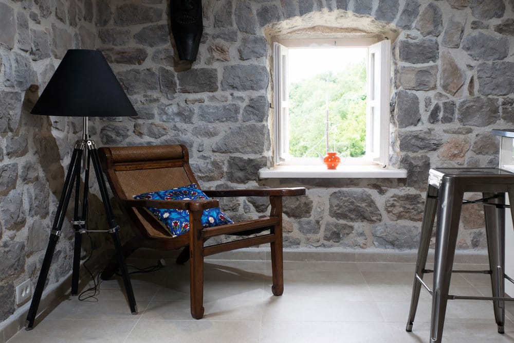 Enterijer dnevne sobe u vili na Luštici, Crna Gora: kameni zidovi, vintage fotelja, podna lampa i pogled kroz prozor na baštu.