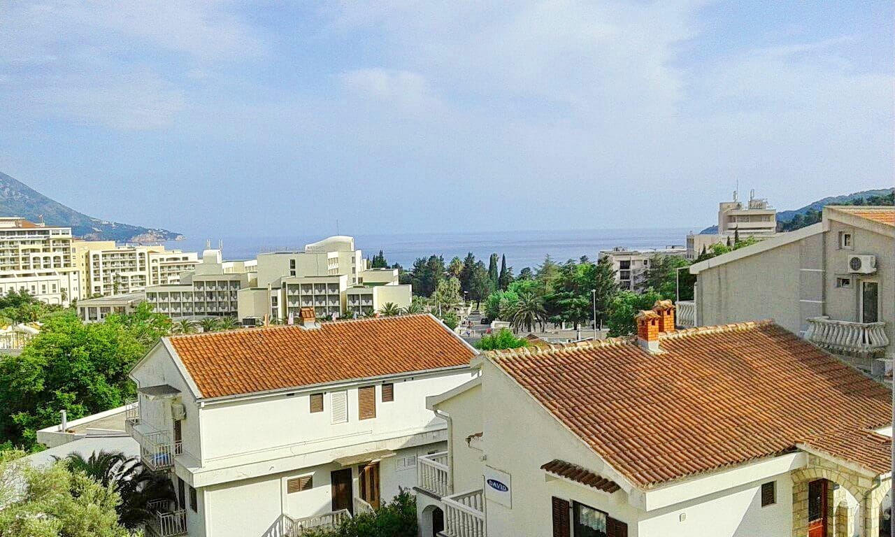 Panoramski pogled na more i letovalište Bečići, Crna Gora, sa terase stana sa krovovima od crepa i zelenim drvećem.