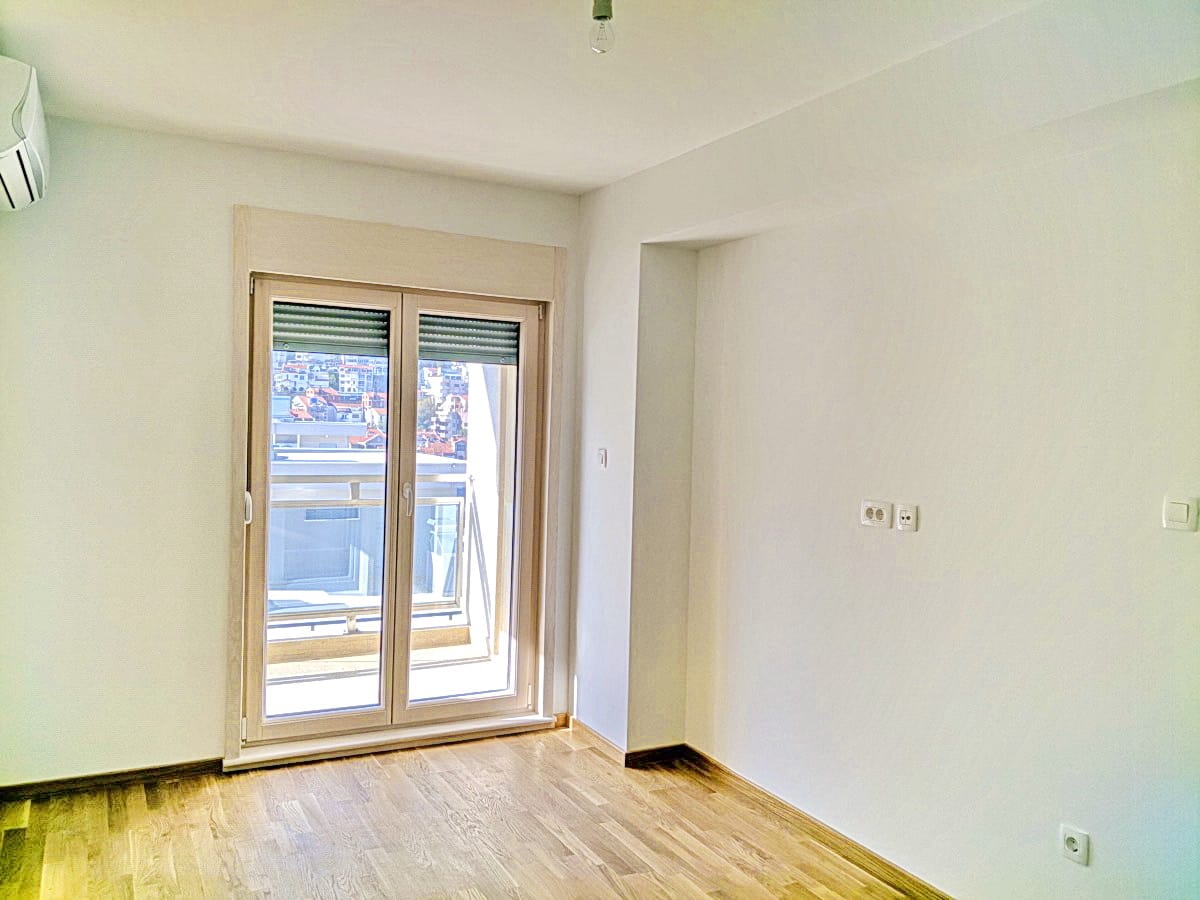 Svetla dnevna soba u stanu u Budvi, Crna Gora: parketni pod, beli zidovi, izlaz na balkon i pogled na grad.