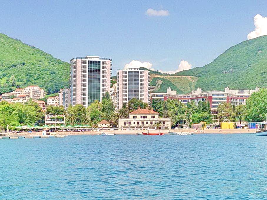 Pogled s mora na moderne stambene komplekse i plažu u Budvi, Crna Gora, u pozadini zelenih planina i letnjeg neba.