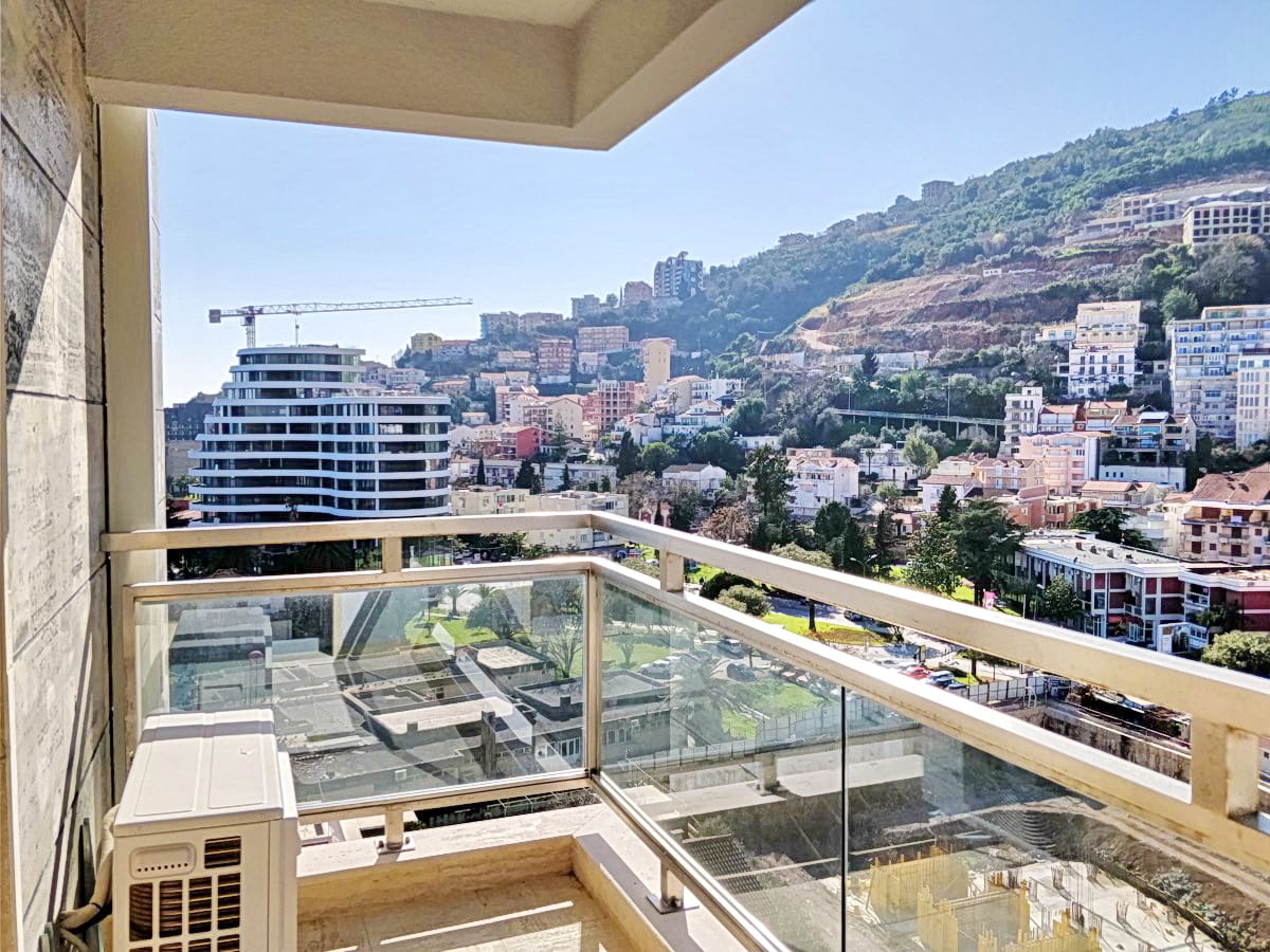 Pogled sa balkona stana u Budvi, Crna Gora: panorama grada, planine i savremena arhitektura sa staklenim fasadama.