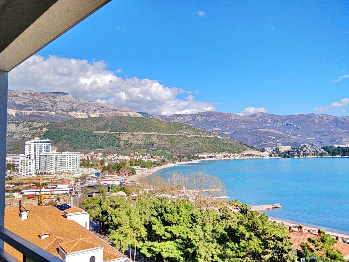 Panoramski pogled sa balkona stana u Budvi, Crna Gora: morska obala, plaža, gradska izgradnja i planinski pejzaž.